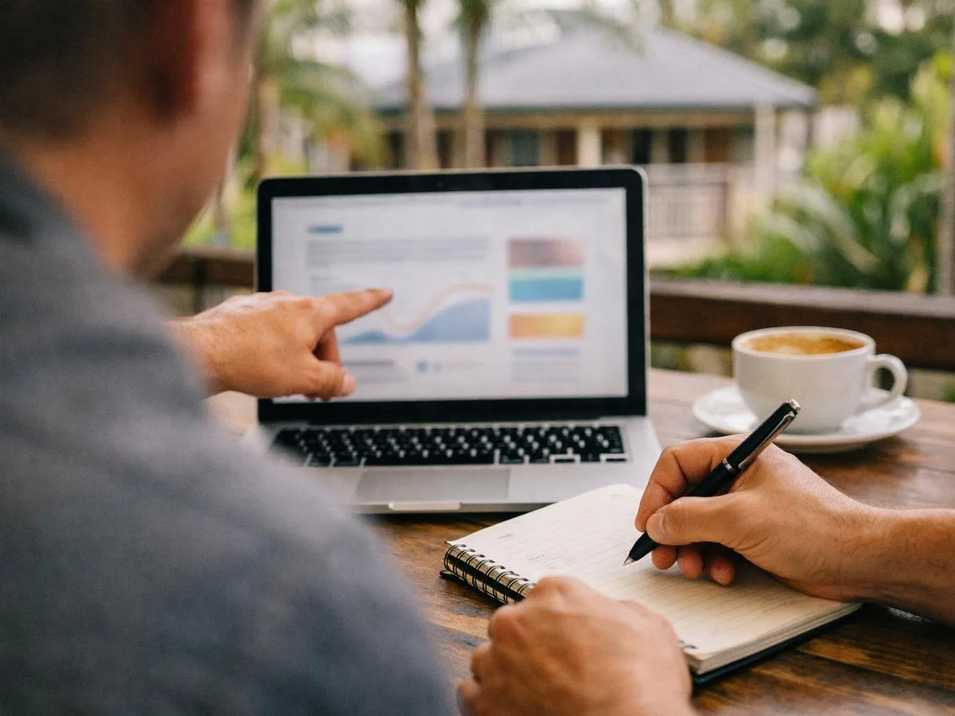 A person points at a laptop chart while writing notes beside a cup of coffee on a wooden table