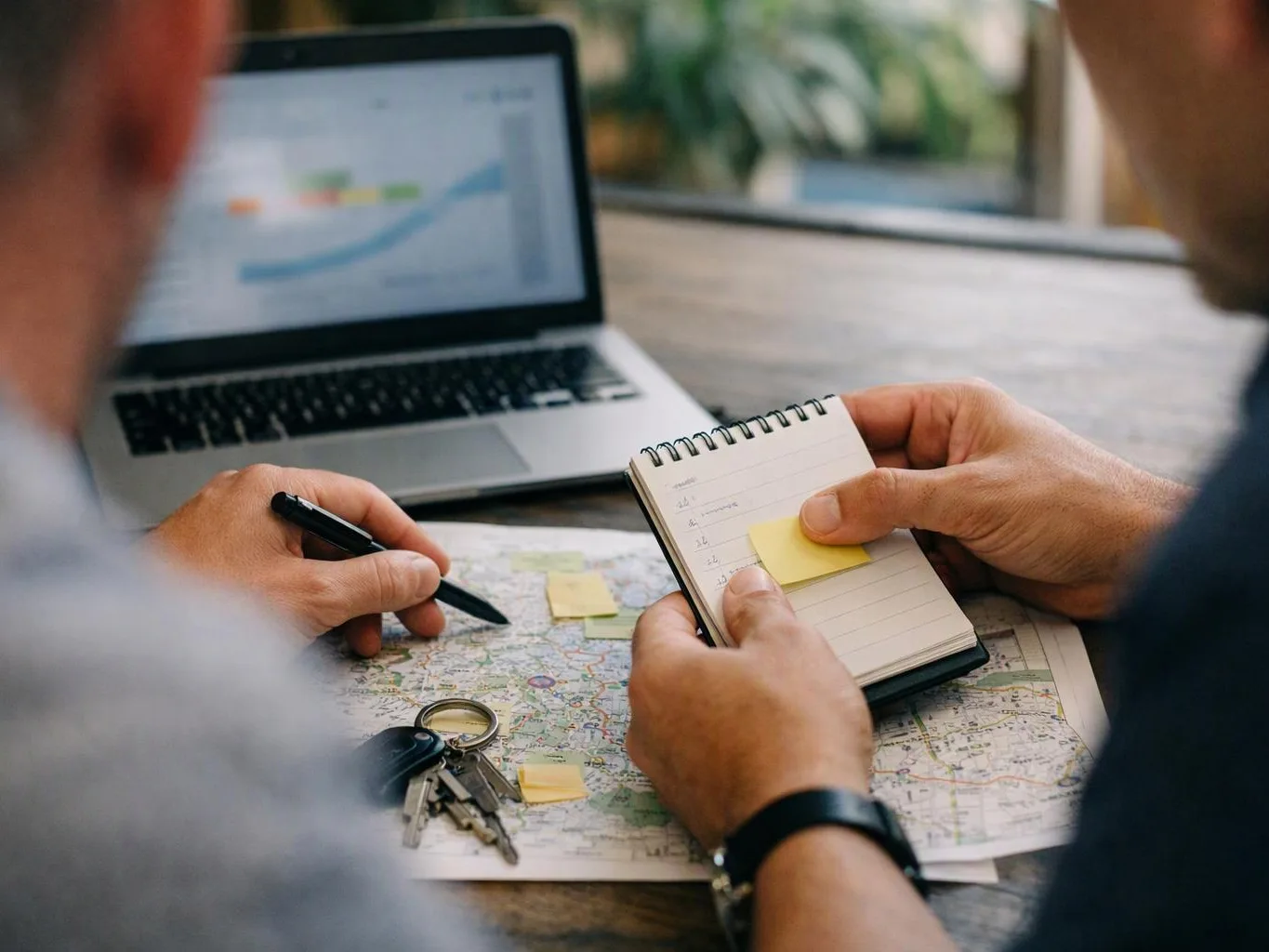 Two people plan a trip at a table, with a map, notebook, keys, and a laptop with a chart
