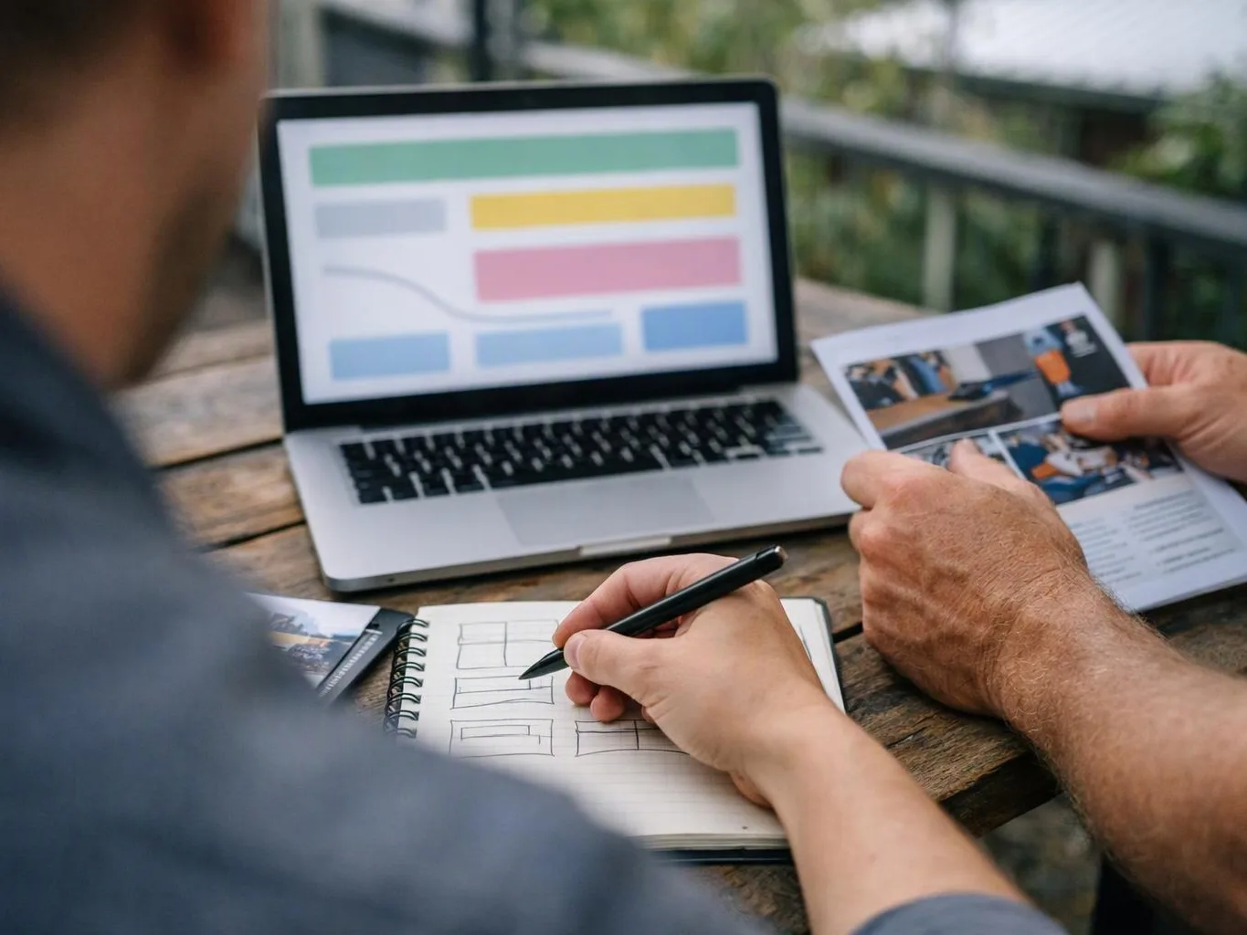 Two people sketch website wireframes beside a laptop and printed mockups at a wooden table